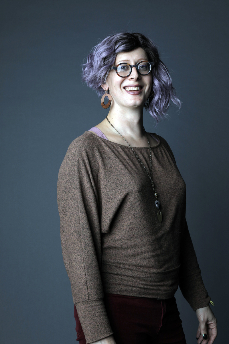 A portrait of Sasha with purple shoulder length hair, brown round glasses, a brown sweater.